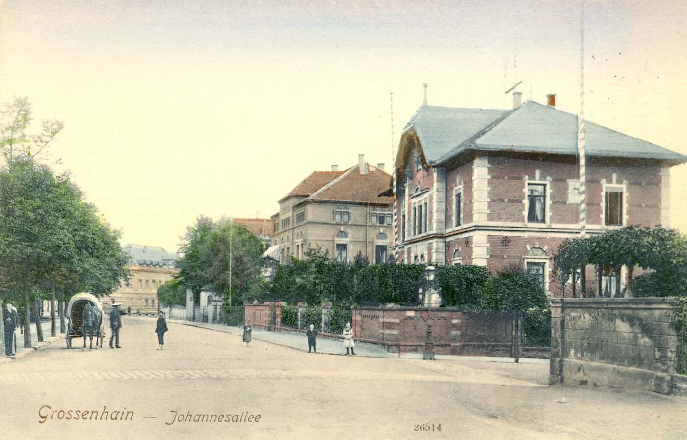 Eine historische, kolorierte Postkarte mit der Beschriftung "Grossenhain - Johannesallee". Zu sehen ist eine breite Straße mit einer Baumreihe, großen Villen, ein Pferdefuhrwerk und wenige Passanten.