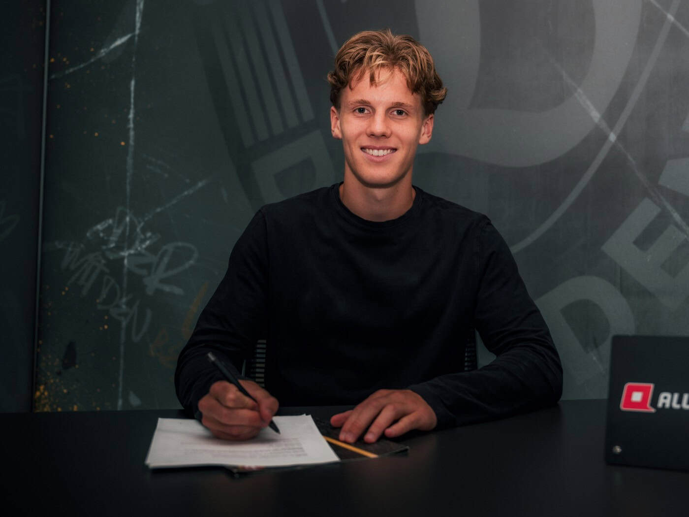 Ein junger, lächelnder Mann mit hellbraunen Locken sitzt an einem schwarzen Tisch und hält einen Stift über ein Papier. Im Hintergrund eine dunkle Wand mit dem Dynamo-Dresden-Logo. Rechts auf dem Tisch steht ein kleiner "ALL-INKL.COM"-Aufsteller.