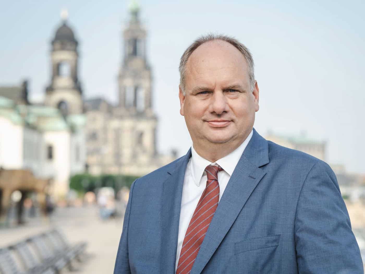 Porträt von Dirk Hilbert im blauen Anzug und mit rot-gestreifter Krawatte. Er blickt lächelnd in die Kamera. Im hellen, leicht unscharfen Hintergrund ist die historische Altstadt von Dresden mit der Katholischen Hofkirche zu sehen.