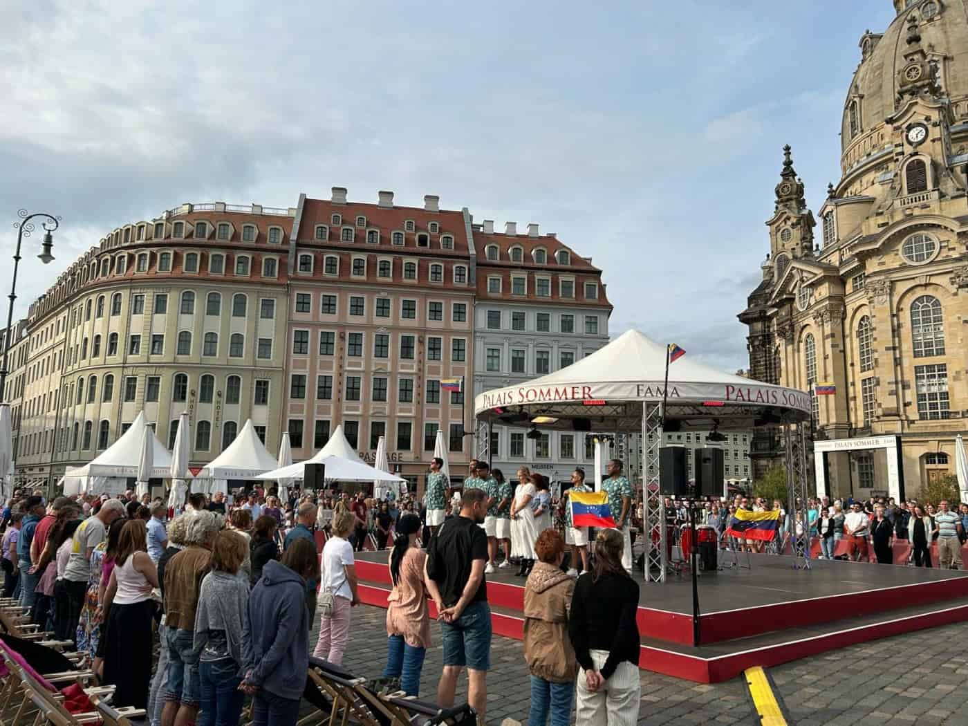Viele Menschen stehen schweigend vor einer überdachten Bühne auf dem Dresdner Neumarkt. Auf der Bühne des Palais Sommer stehen Künstler mit einer venezolanischen Flagge. Rechts im Hintergrund ragt die Kuppel der Frauenkirche auf.