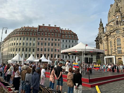 Viele Menschen stehen schweigend vor einer überdachten Bühne auf dem Dresdner Neumarkt. Auf der Bühne des Palais Sommer stehen Künstler mit einer venezolanischen Flagge. Rechts im Hintergrund ragt die Kuppel der Frauenkirche auf.