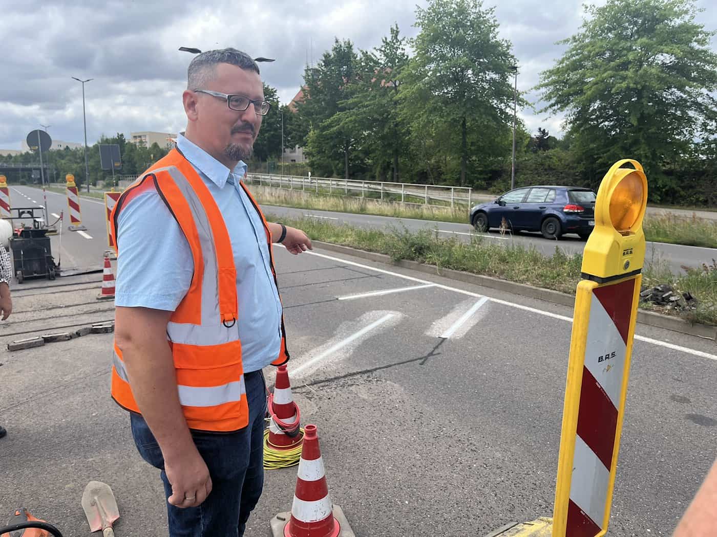 Ein Mann im hellblauen Hemd und einer orangefarbenen Warnweste zeigt auf Fahrbahnmarkierungen auf dem Asphalt. Im Hintergrund fährt ein blaues Auto vorbei. Rechts steht eine rot-weiß gestreifte Absperrung mit gelber Leuchte.