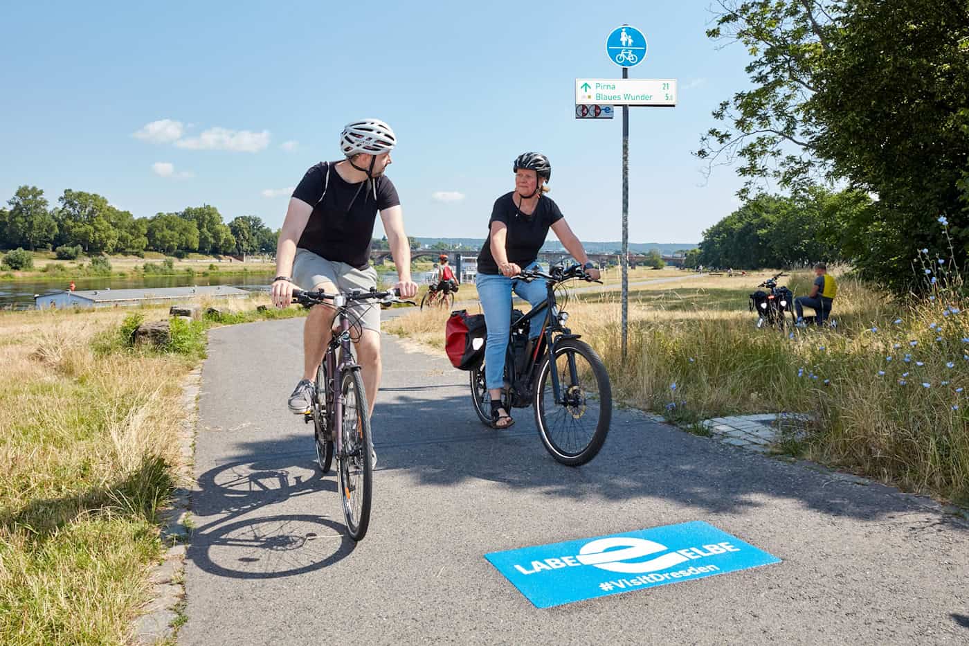 Ein Mann und eine Frau fahren auf Fahrrädern mit Helmen den Elberadweg entlang. Auf dem Asphalt vor ihnen befindet sich ein blaues Piktogramm mit der Aufschrift Labe Elbe und dem Hashtag VisitDresden.