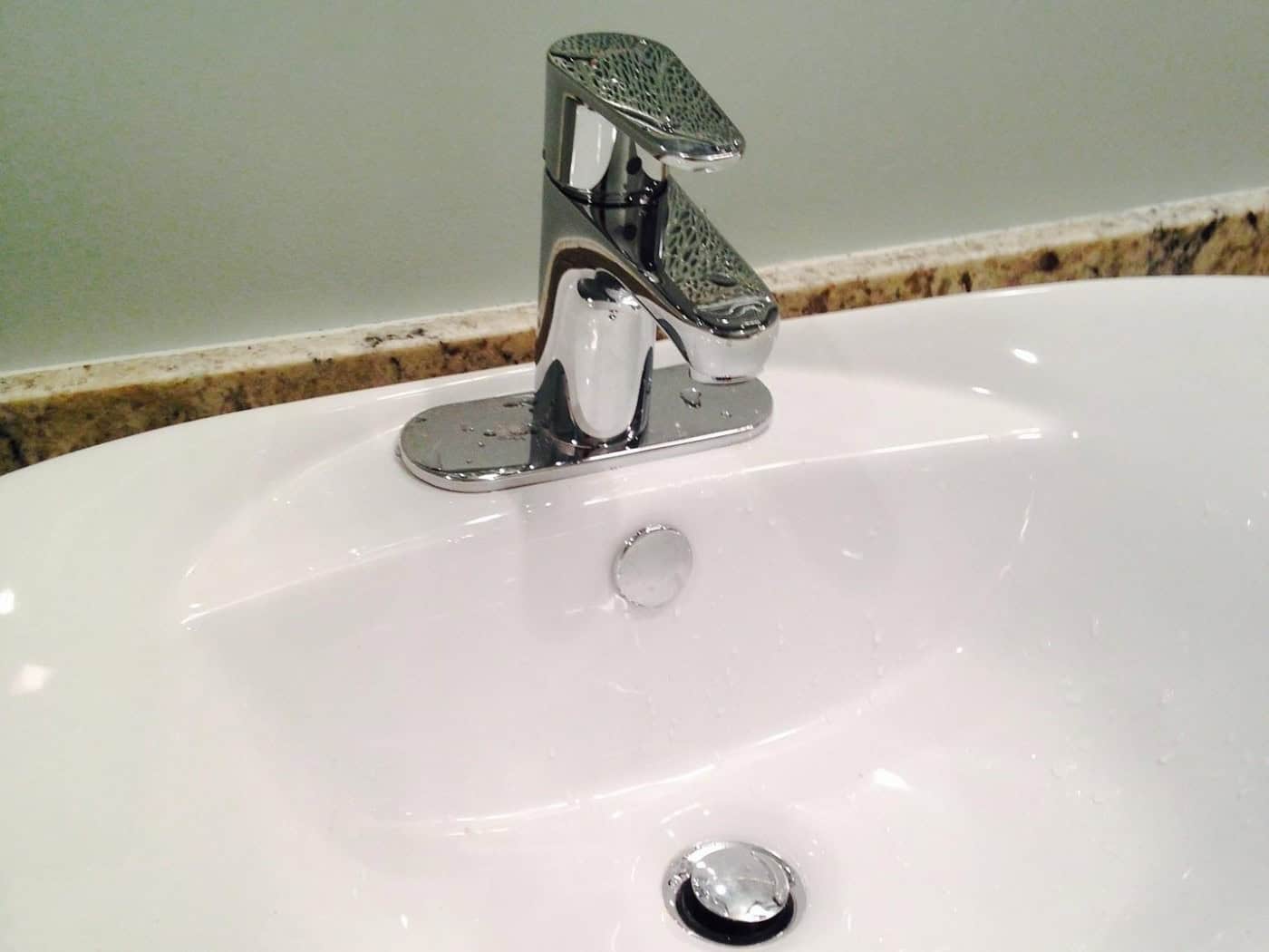 Nahaufnahme eines modernen, weißen Keramik-Waschbeckens im Badezimmer. Auf dem Becken ist eine glänzende, geschlossene verchromte Mischbatterie montiert. Das Waschbecken steht auf einer gemusterten Granit-Arbeitsplatte vor einer hellgrünen Wand.