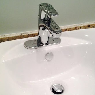 Nahaufnahme eines modernen, weißen Keramik-Waschbeckens im Badezimmer. Auf dem Becken ist eine glänzende, geschlossene verchromte Mischbatterie montiert. Das Waschbecken steht auf einer gemusterten Granit-Arbeitsplatte vor einer hellgrünen Wand.