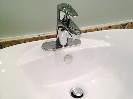 Nahaufnahme eines modernen, weißen Keramik-Waschbeckens im Badezimmer. Auf dem Becken ist eine glänzende, geschlossene verchromte Mischbatterie montiert. Das Waschbecken steht auf einer gemusterten Granit-Arbeitsplatte vor einer hellgrünen Wand.