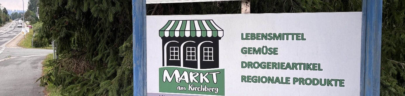 Ein Schild am Straßenrand mit dem Text "Markt am Kirchberg: Lebensmittel, Gemüse, Drogerieartikel, Regionale Produkte".