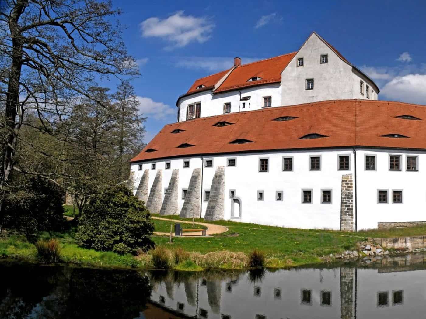 Ein Schloss, das eher massiv wie eine Burg gebaut ist, mit weißer Fassade und rotem Dach. Davor ein Gewässer. Zum Schlosstor hin eine Brücke.