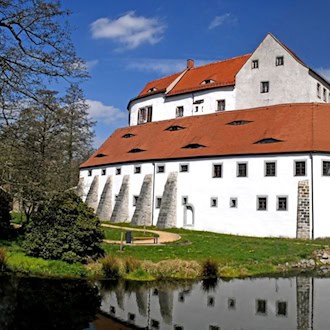 Ein Schloss, das eher massiv wie eine Burg gebaut ist, mit weißer Fassade und rotem Dach. Davor ein Gewässer. Zum Schlosstor hin eine Brücke.