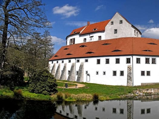Ein Schloss, das eher massiv wie eine Burg gebaut ist, mit weißer Fassade und rotem Dach. Davor ein Gewässer. Zum Schlosstor hin eine Brücke.