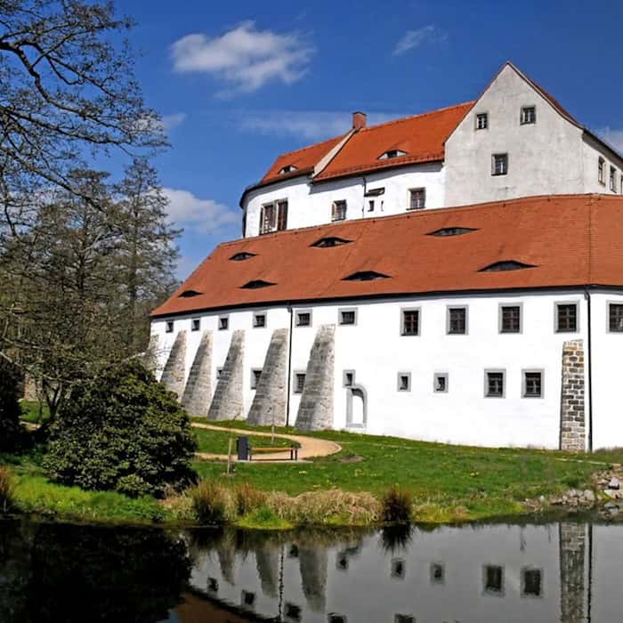 Ein Schloss, das eher massiv wie eine Burg gebaut ist, mit weißer Fassade und rotem Dach. Davor ein Gewässer. Zum Schlosstor hin eine Brücke.