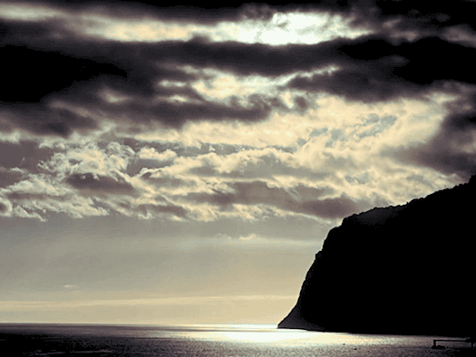 Eine dramatische Meereslandschaft unter einem dunklen, dicht bewölkten Himmel, durch den helles Sonnenlicht bricht und auf dem Wasser glänzt. Rechts ragt die tiefschwarze Silhouette einer steilen Felsküste ins Bild.