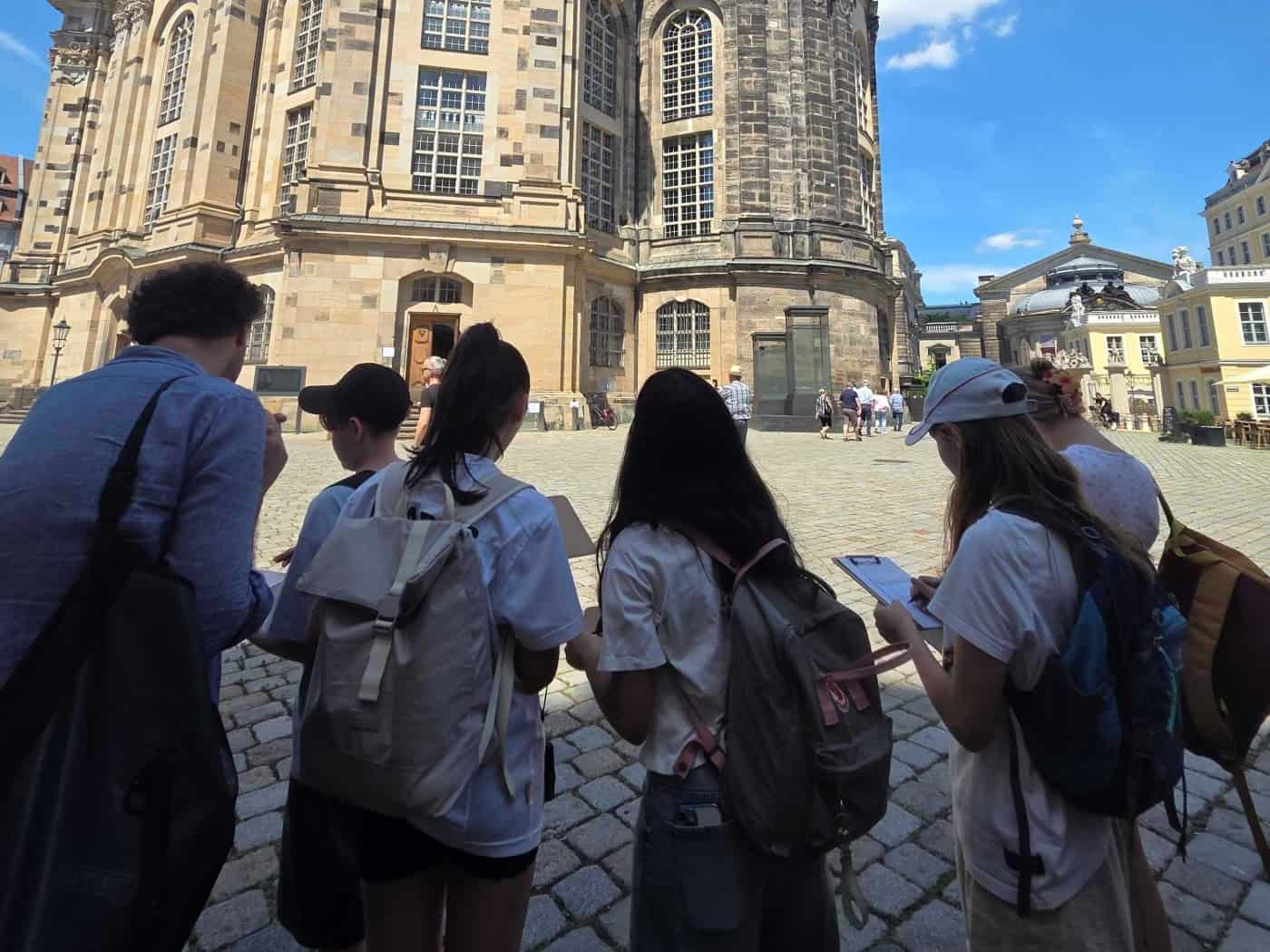 Eine Gruppe von Jugendlichen mit Rucksäcken steht von hinten gesehen auf dem Pflaster des Dresdner Neumarkts und blickt hoch zur sandsteinfarbenen Frauenkirche. Zwei Mädchen schreiben auf Klemmbretter. Im Hintergrund ist ein strahlend blauer Himmel mit hellen Wolken.