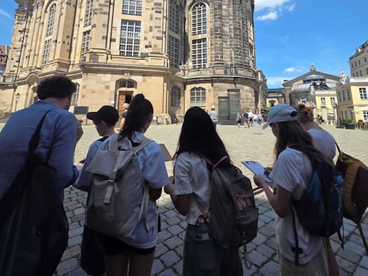 Eine Gruppe von Jugendlichen mit Rucksäcken steht von hinten gesehen auf dem Pflaster des Dresdner Neumarkts und blickt hoch zur sandsteinfarbenen Frauenkirche. Zwei Mädchen schreiben auf Klemmbretter. Im Hintergrund ist ein strahlend blauer Himmel mit hellen Wolken.