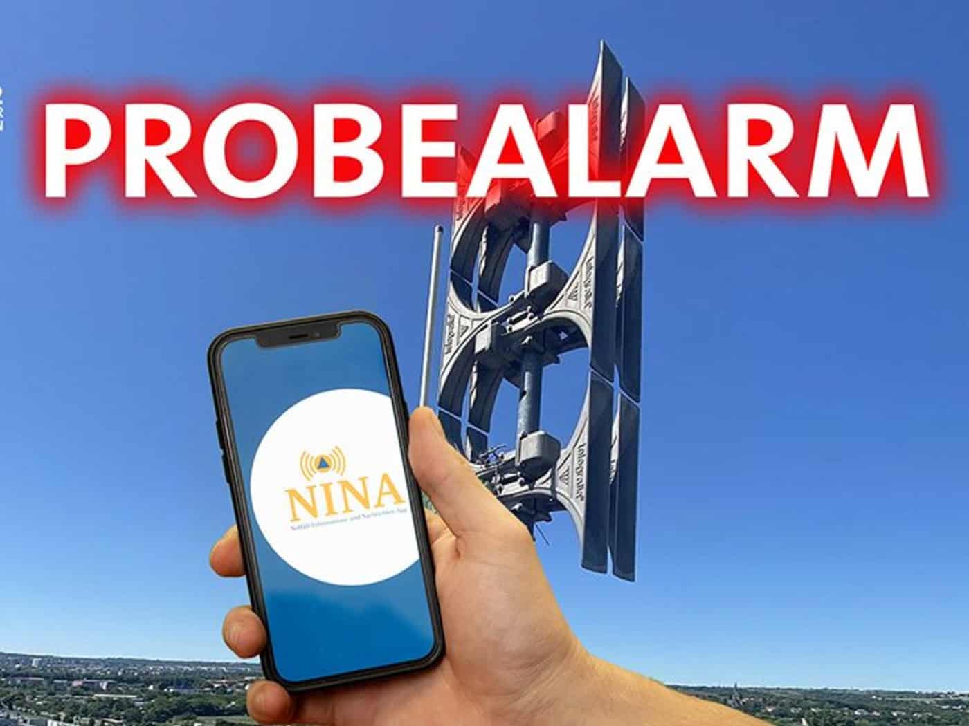 Eine Hand hält ein Smartphone mit dem runden, weißen Logo der NINA-App. Im Hintergrund steht eine graue elektronische Sirenenanlage vor blauem Himmel über einer Stadt. Oben leuchtet das rote Wort „PROBEALARM“.