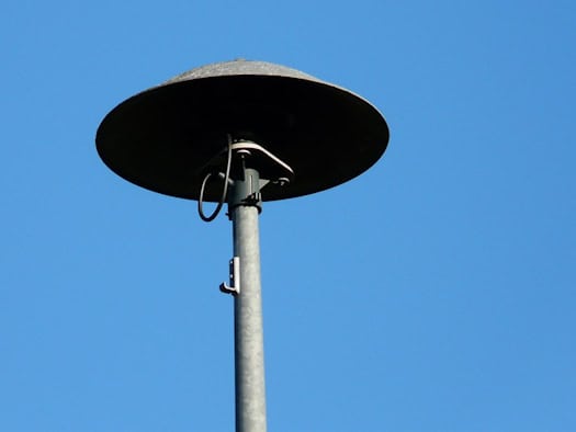 Eine pilzförmige, dunkle Sirene ist auf einem runden Metallmast montiert. Sie hebt sich deutlich vor einem vollkommen wolkenlosen, strahlend blauen Himmel ab. Das Bild zeigt die Anlage in einer Nahaufnahme von unten bei klarem Tageslicht.