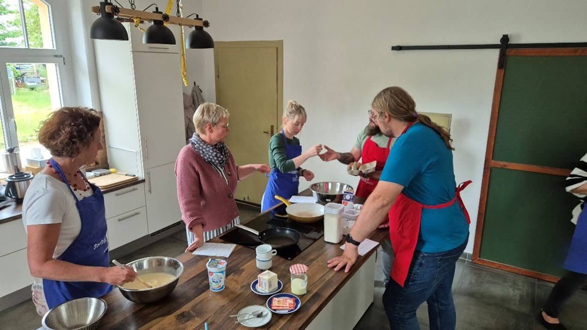 Mehrere Personen in einer Küche bereiten gemeinsam etwas zu. Sie tragen Schürzen und stehen um eine Arbeitsfläche mit Kochutensilien, Zutaten und einem Kochfeld.