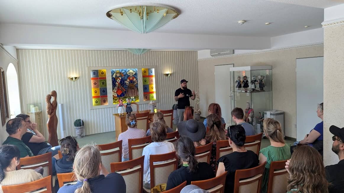 Ein Mann hält einen Vortrag vor einem sitzenden Publikum, im Hintergrund Puppen und Abbildungen mit sorbischer Tracht