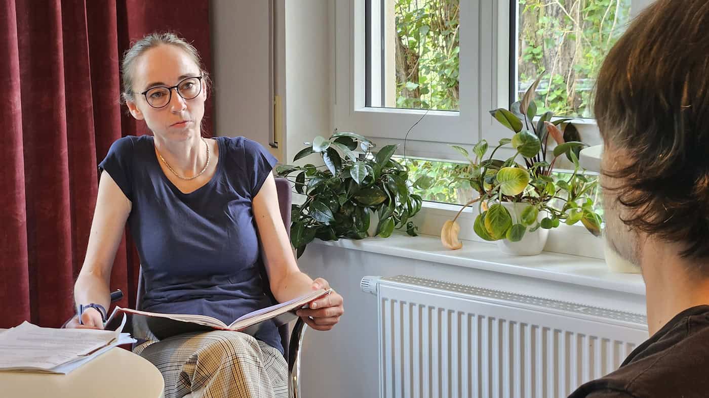 Sozialbürgermeisterin Kaufmann zu Besuch in der AWO-Schuldnerberatung Dresden-Pieschen Eine Frau mit Brille und dunkelblauem T-Shirt sitzt mit einem offenen Notizbuch an einem Tisch und spricht mit einem Mann, der im Vordergrund von hinten zu sehen ist. Im Hintergrund stehen Zimmerpflanzen auf einer Fensterbank vor einem Fenster.