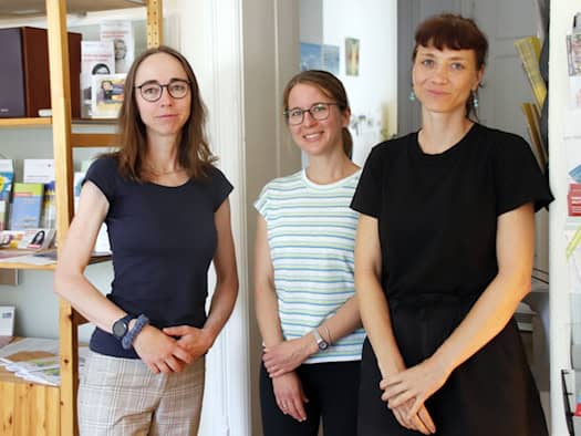Drei Frauen stehen lächelnd nebeneinander in einem Raum. Links steht Bürgermeisterin Kaufmann mit verschränkten Händen, daneben zwei Mitarbeiterinnen im gestreiften und schwarzen T-Shirt. Links ist ein Holzregal mit Flyern und einer Stereoanlage.