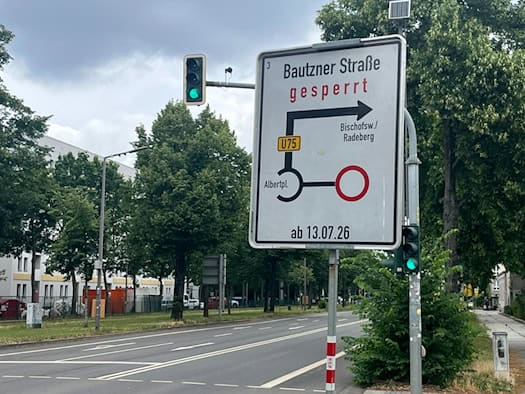 Ein großes weißes Hinweisschild an einer Straße mit der Aufschrift „Bautzner Straße gesperrt ab 13.07.26“ und einer gezeichneten Umleitungsstrecke für die U75. Im Hintergrund stehen grüne Bäume und Wohngebäude unter einem bewölkten Himmel.