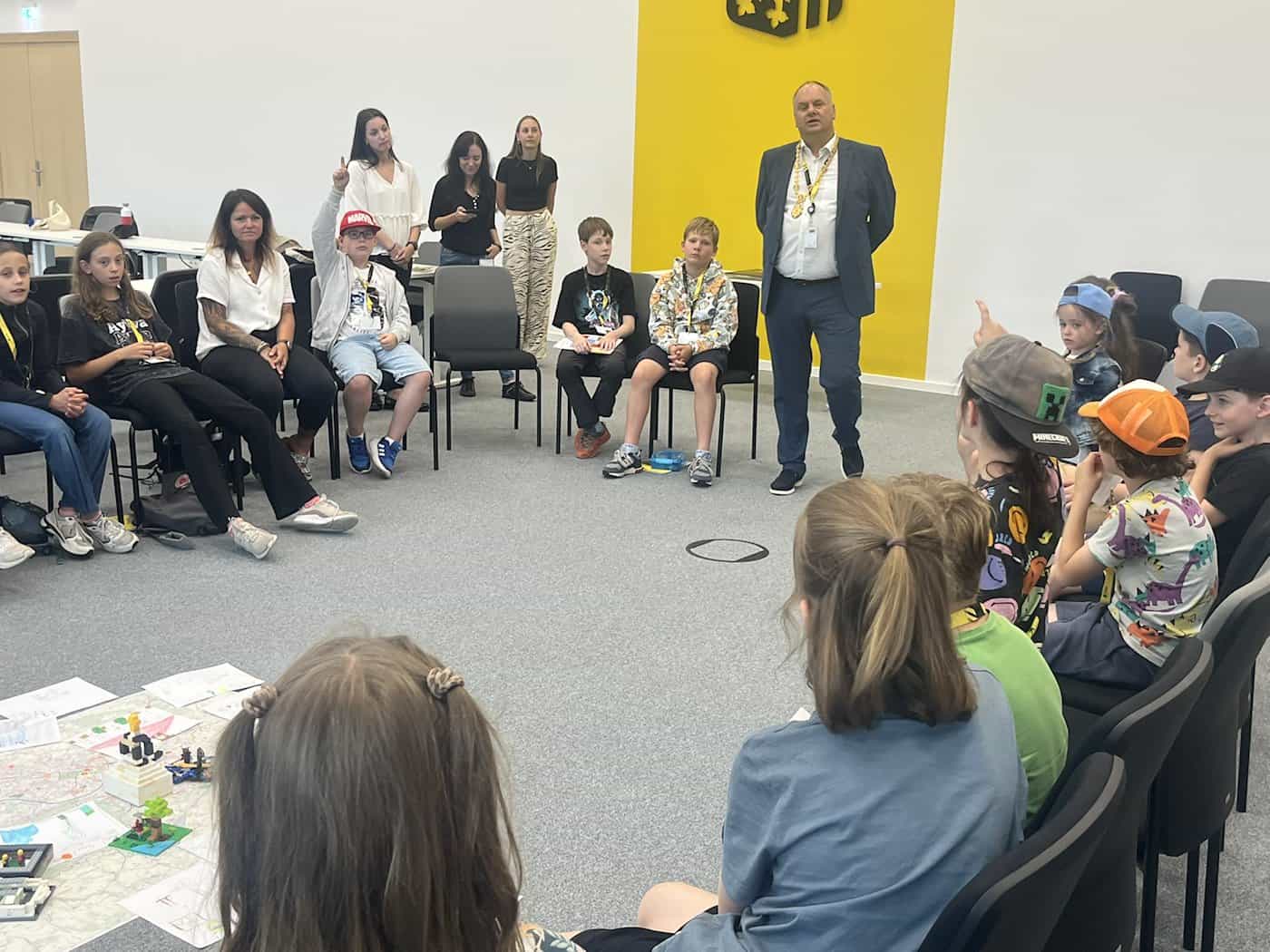 Großer Stuhlkreis im Stadtforum: Kinder und Betreuer sitzen um einen Stadtplan mit Lego-Modellen auf dem Boden. Im Hintergrund steht OB Dirk Hilbert mit goldener Amtskette vor einer gelben Wand.