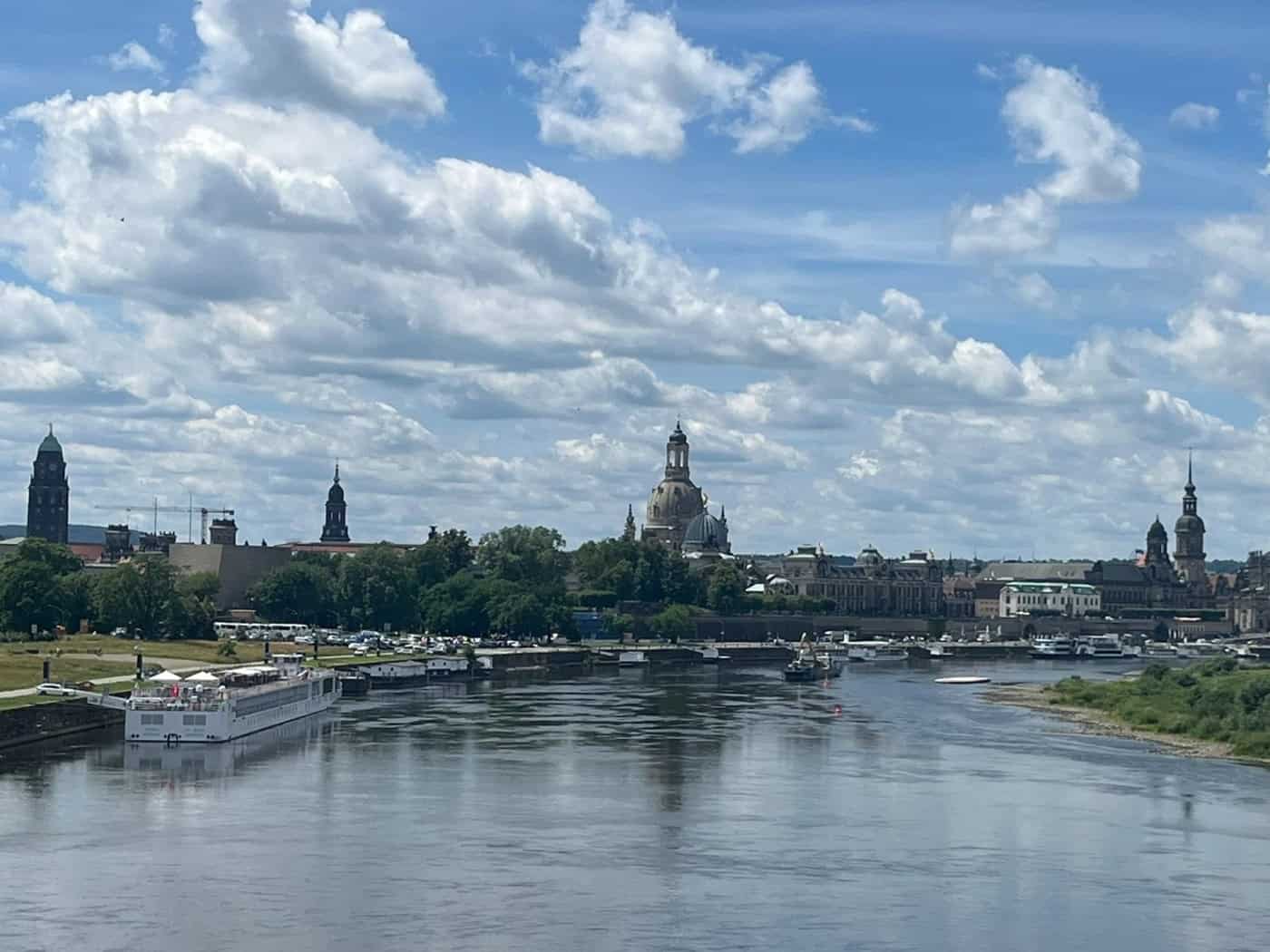 Weiter Blick über die Elbe auf die historische Silhouette Dresdens mit der Frauenkirche. Im Vordergrund fließt der Fluss an der Stelle der ehemaligen Carolabrücke. Links liegen Schiffe am Ufer, rechts erstrecken sich grüne Wiesen unter einem bewölkten Himmel.