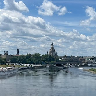 Weiter Blick über die Elbe auf die historische Silhouette Dresdens mit der Frauenkirche. Im Vordergrund fließt der Fluss an der Stelle der ehemaligen Carolabrücke. Links liegen Schiffe am Ufer, rechts erstrecken sich grüne Wiesen unter einem bewölkten Himmel.