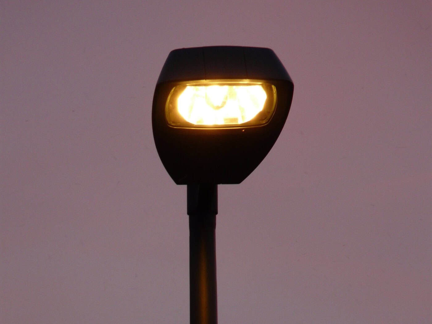 Nahaufnahme einer modernen, dunklen Straßenlaterne, die mit einem warmen, gelben Licht leuchtet. Das Gehäuse der Lampe hebt sich kontrastreich von einem min