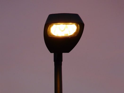Nahaufnahme einer modernen, dunklen Straßenlaterne, die mit einem warmen, gelben Licht leuchtet. Das Gehäuse der Lampe hebt sich kontrastreich von einem min