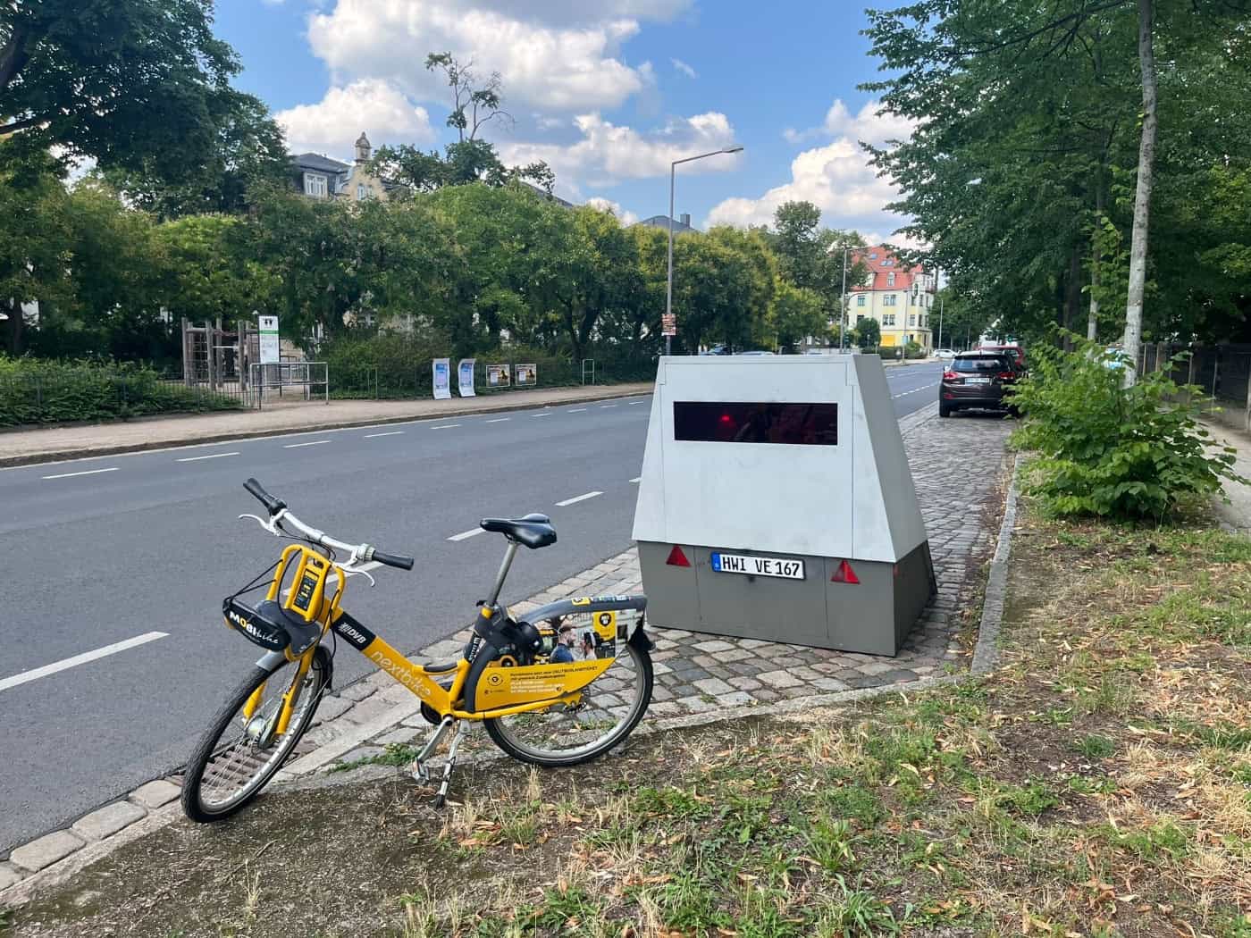 Ein gelbes Leihfahrrad parkt auf einem Grünstreifen im Vordergrund. Dahinter steht ein grauer, kastenförmiger Blitzer-Messanhänger mit dem Kennzeichen HWI VE 167 am Rand einer Straße in Dresden. Im Hintergrund sind grüne Bäume und ein Spielplatz zu sehen.