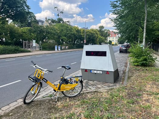 Ein gelbes Leihfahrrad parkt auf einem Grünstreifen im Vordergrund. Dahinter steht ein grauer, kastenförmiger Blitzer-Messanhänger mit dem Kennzeichen HWI VE 167 am Rand einer Straße in Dresden. Im Hintergrund sind grüne Bäume und ein Spielplatz zu sehen.