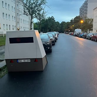Ein grauer Blitzer-Anhänger mit dem Kennzeichen HWI VE 159 steht am linken Rand einer nassen Straße bei Dämmerung. Links und rechts befinden sich mehrstöckige Wohnhäuser, vor dem Anhänger parken Autos. Am Himmel hängen dunkle Wolken.