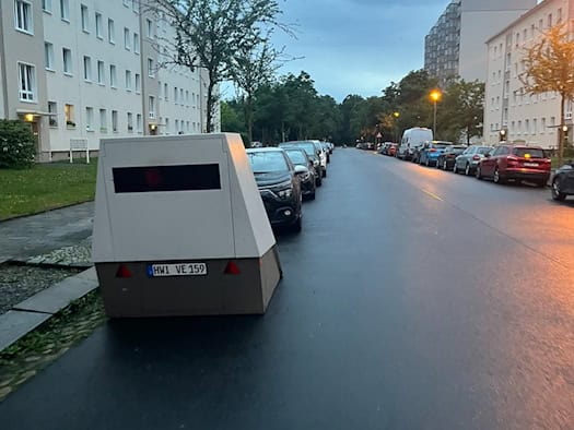 Ein grauer Blitzer-Anhänger mit dem Kennzeichen HWI VE 159 steht am linken Rand einer nassen Straße bei Dämmerung. Links und rechts befinden sich mehrstöckige Wohnhäuser, vor dem Anhänger parken Autos. Am Himmel hängen dunkle Wolken.