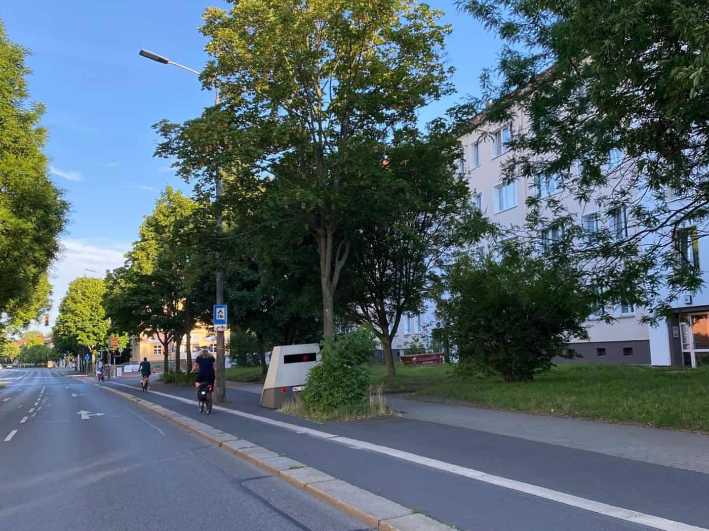 Ein grauer Blitzer-Anhänger steht getarnt hinter einem kleinen grünen Busch am Rand einer mehrspurigen Straße neben einem Fahrradweg, auf dem ein Radfahrer fährt. Im Hintergrund sind Bäume, ein helles Wohnhaus und ein wolkenloser blauer Himmel zu sehen.