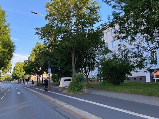 Ein grauer Blitzer-Anhänger steht getarnt hinter einem kleinen grünen Busch am Rand einer mehrspurigen Straße neben einem Fahrradweg, auf dem ein Radfahrer fährt. Im Hintergrund sind Bäume, ein helles Wohnhaus und ein wolkenloser blauer Himmel zu sehen.