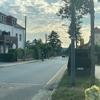 Ein dunkelgrüner, kastenförmiger Blitzer-Anhänger mit dem Kennzeichen HWI-VE 213 steht am rechten Rand einer Wohnstraße hinter einem Baum. Links sind helle Wohnhäuser zu sehen, im Vordergrund auf dem Grünstreifen liegt ein prall gefüllter gelber Sack.