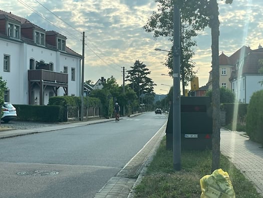 Ein dunkelgrüner, kastenförmiger Blitzer-Anhänger mit dem Kennzeichen HWI-VE 213 steht am rechten Rand einer Wohnstraße hinter einem Baum. Links sind helle Wohnhäuser zu sehen, im Vordergrund auf dem Grünstreifen liegt ein prall gefüllter gelber Sack.