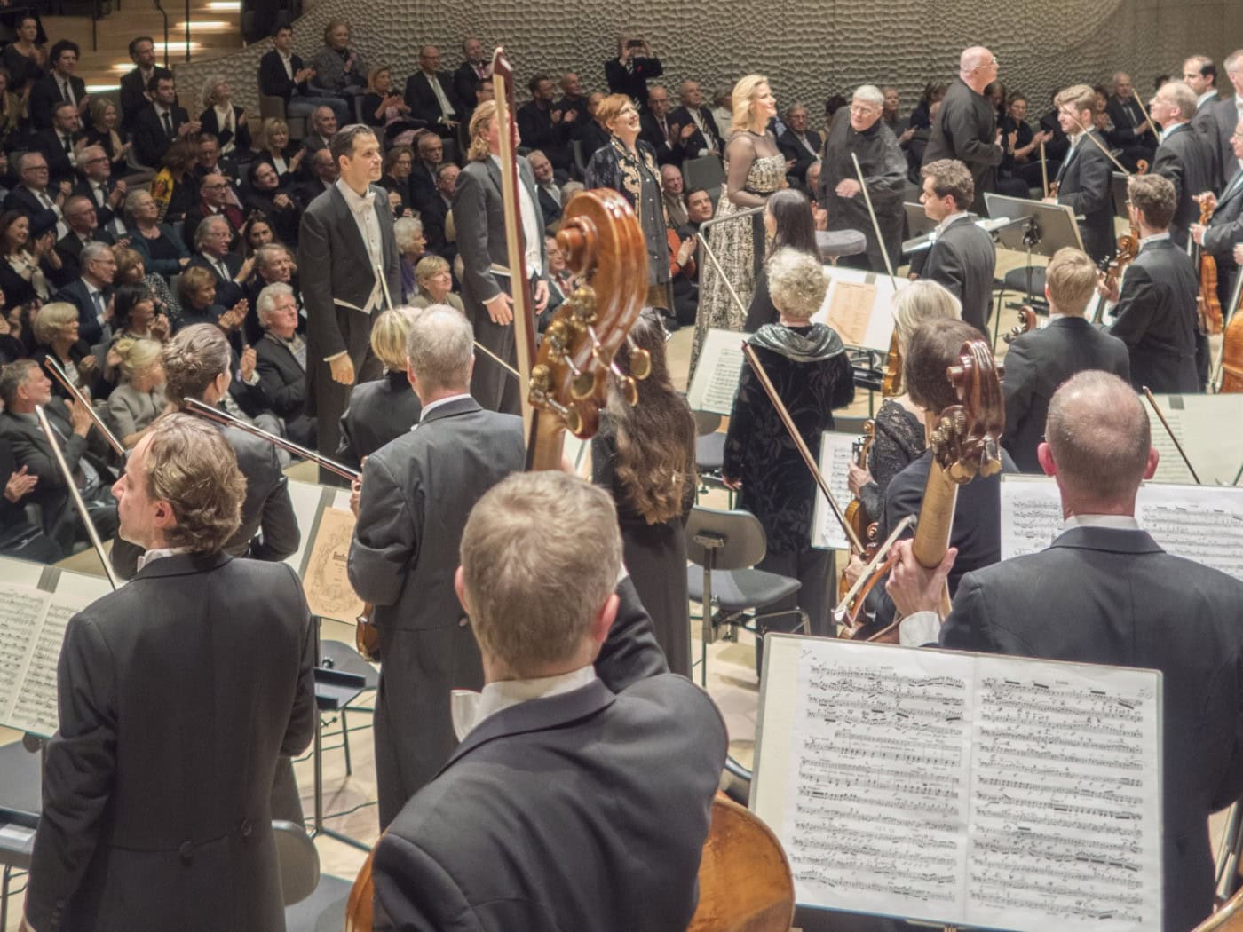 Blick von der Bühne auf einen modernen, hellen Konzertsaal mit geschwungenen Rängen, die voll besetzt mit applaudierenden Zuschauern sind. Im Vordergrund stehen Musiker eines Orchesters und Solisten mit Notenständern auf der Bühne.