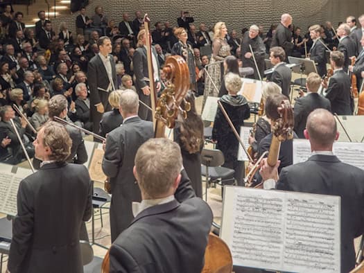 Blick von der Bühne auf einen modernen, hellen Konzertsaal mit geschwungenen Rängen, die voll besetzt mit applaudierenden Zuschauern sind. Im Vordergrund stehen Musiker eines Orchesters und Solisten mit Notenständern auf der Bühne.