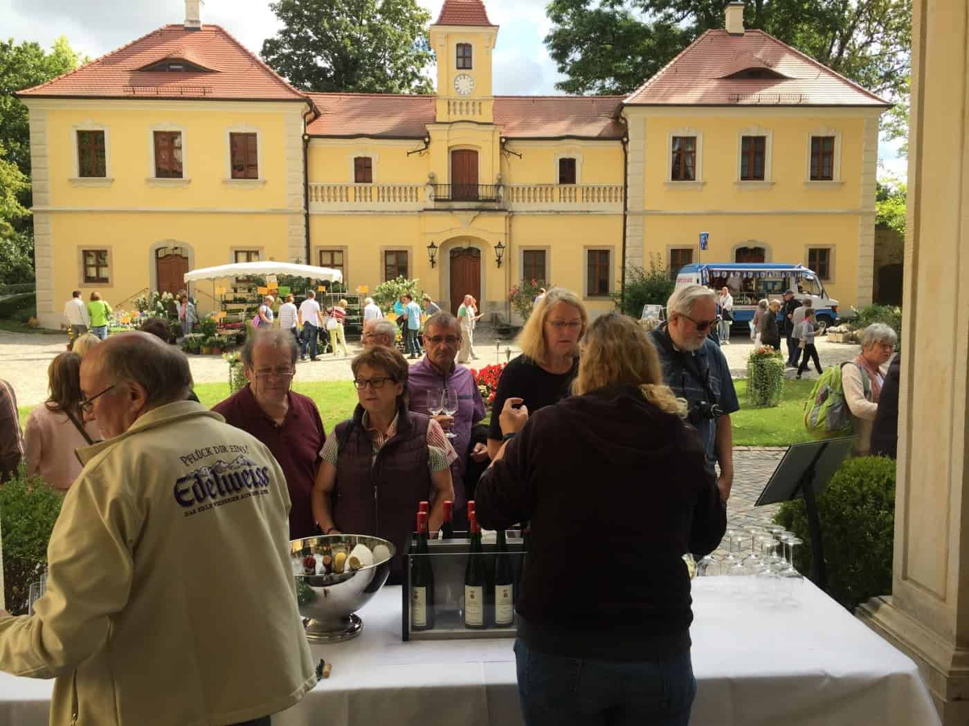 Mehrere Besucher stehen an einem weißen Tisch mit Weinflaschen und Gläsern im Hof von Schloss Proschwitz. Im Hintergrund strahlt das zweistöckige, gelbe Barockschloss mit rotem Dach unter leicht bewölktem Himmel. Im Garten stehen weitere Marktstände.