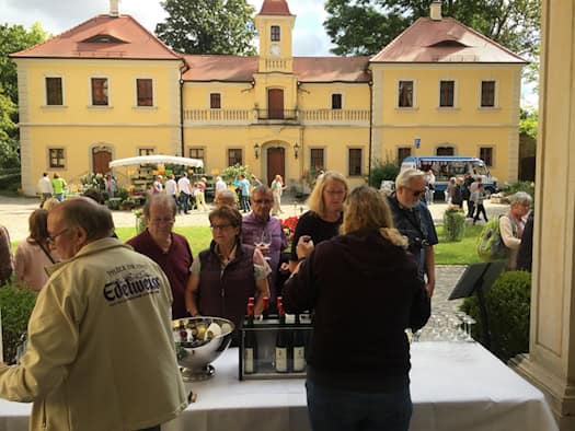 Mehrere Besucher stehen an einem weißen Tisch mit Weinflaschen und Gläsern im Hof von Schloss Proschwitz. Im Hintergrund strahlt das zweistöckige, gelbe Barockschloss mit rotem Dach unter leicht bewölktem Himmel. Im Garten stehen weitere Marktstände.