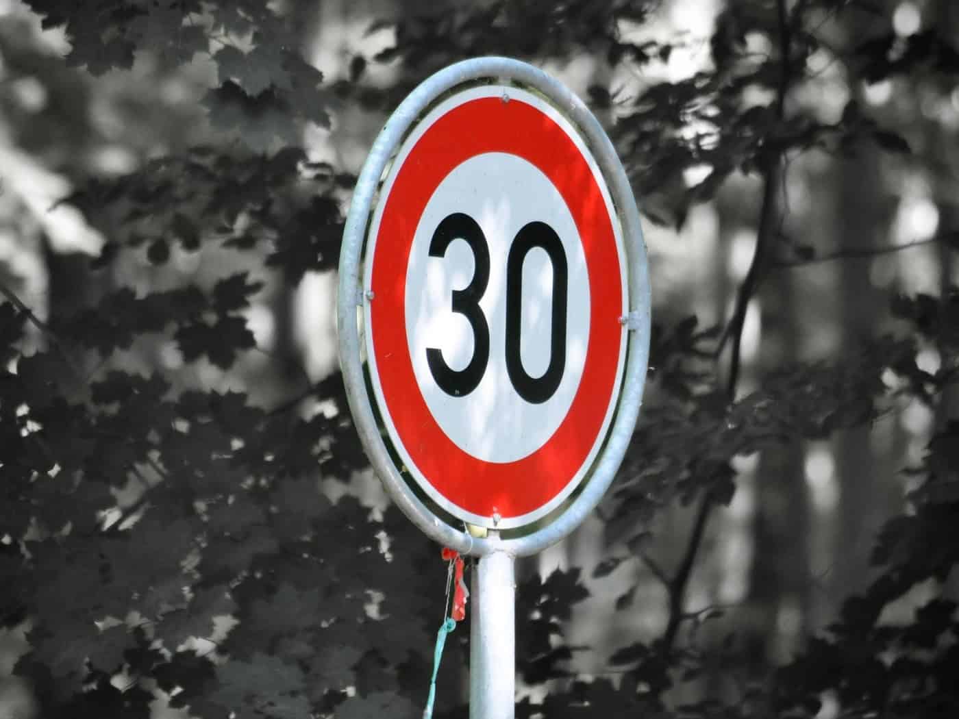 Ein rundes Verkehrsschild mit rotem Rand und weißem Grund zeigt die schwarze Zahl 30. Das Schild steht farbig im Fokus vor einem dicht bewachsenen, verschwommenen Hintergrund aus Blättern und Bäumen in Schwarz-Weiß-Optik.