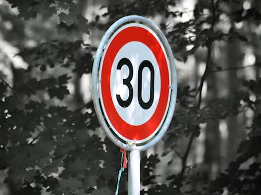 Ein rundes Verkehrsschild mit rotem Rand und weißem Grund zeigt die schwarze Zahl 30. Das Schild steht farbig im Fokus vor einem dicht bewachsenen, verschwommenen Hintergrund aus Blättern und Bäumen in Schwarz-Weiß-Optik.