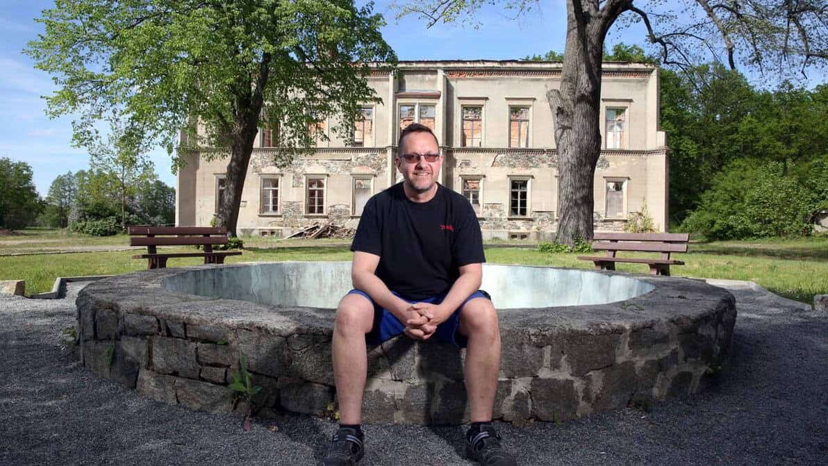 Ein Mann in kurzer Hose und T-Shirt sitzt auf dem Rand eines gemauerten Brunnens und blickt lächelnd in die Kamera. Im Hintergrund ein Herrenhaus bzw. Schloss mit sichtbaren baulichen Schäden.
