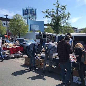 Besucher stöbern an Trödelständen auf einem sonnigen Parkplatz. Im Hintergrund ist das markante blaue Gebäude des Elbeparks Dresden mit großem Logo zu sehen. Weiße Transporter stehen neben Verkaufstischen mit Kisten, Geschirr und Büchern.