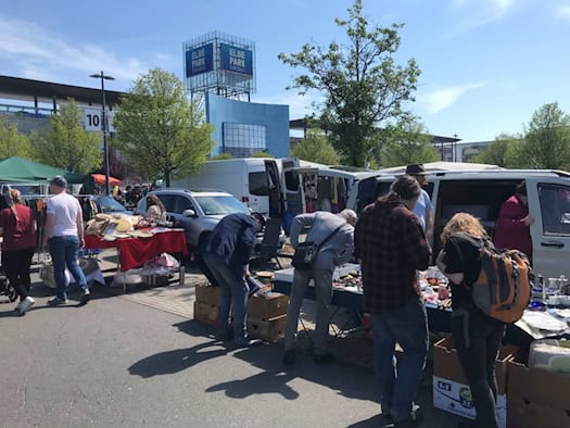Besucher stöbern an Trödelständen auf einem sonnigen Parkplatz. Im Hintergrund ist das markante blaue Gebäude des Elbeparks Dresden mit großem Logo zu sehen. Weiße Transporter stehen neben Verkaufstischen mit Kisten, Geschirr und Büchern.