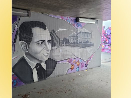 Ein Grafitto auf einer Betonwand in einer Unterführung. Abgebildet ist der Kopf eines Mannes im Anzug sowie eine Straßenansicht mit einer Baumreihe und Villen.