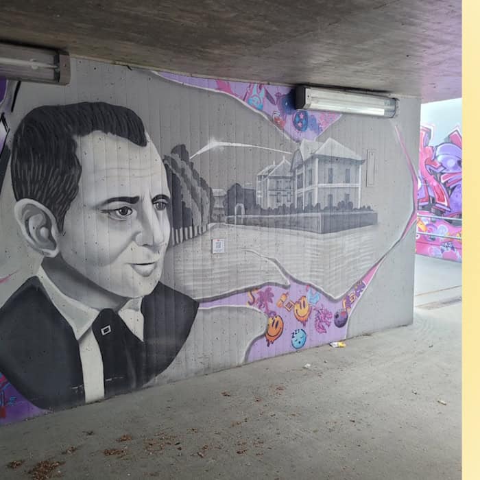 Ein Grafitto auf einer Betonwand in einer Unterführung. Abgebildet ist der Kopf eines Mannes im Anzug sowie eine Straßenansicht mit einer Baumreihe und Villen.