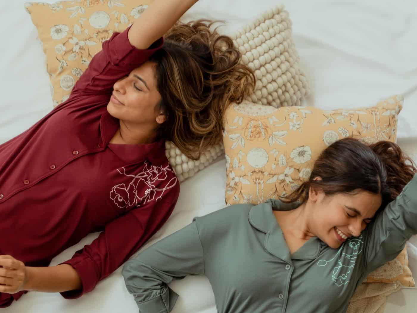 Zwei Frauen in weinroten und olivgrünen Pyjamas liegen lachend auf hellen Kissen. Aus der Vogelperspektive ist eine gemütliche Decke zu sehen.